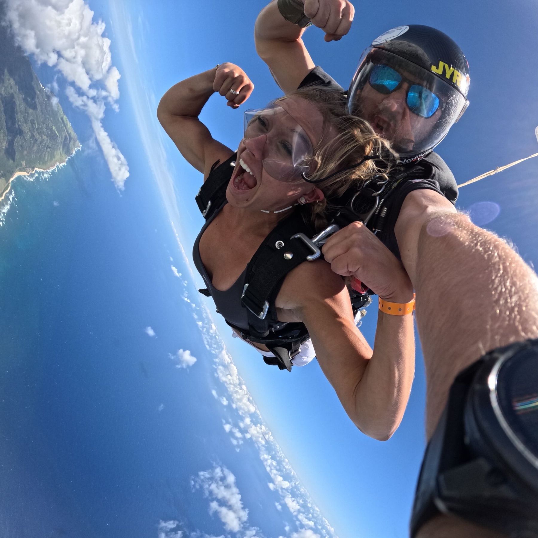 The coach tandem skydiving over a coastline, smiling wide and flexing one arm.