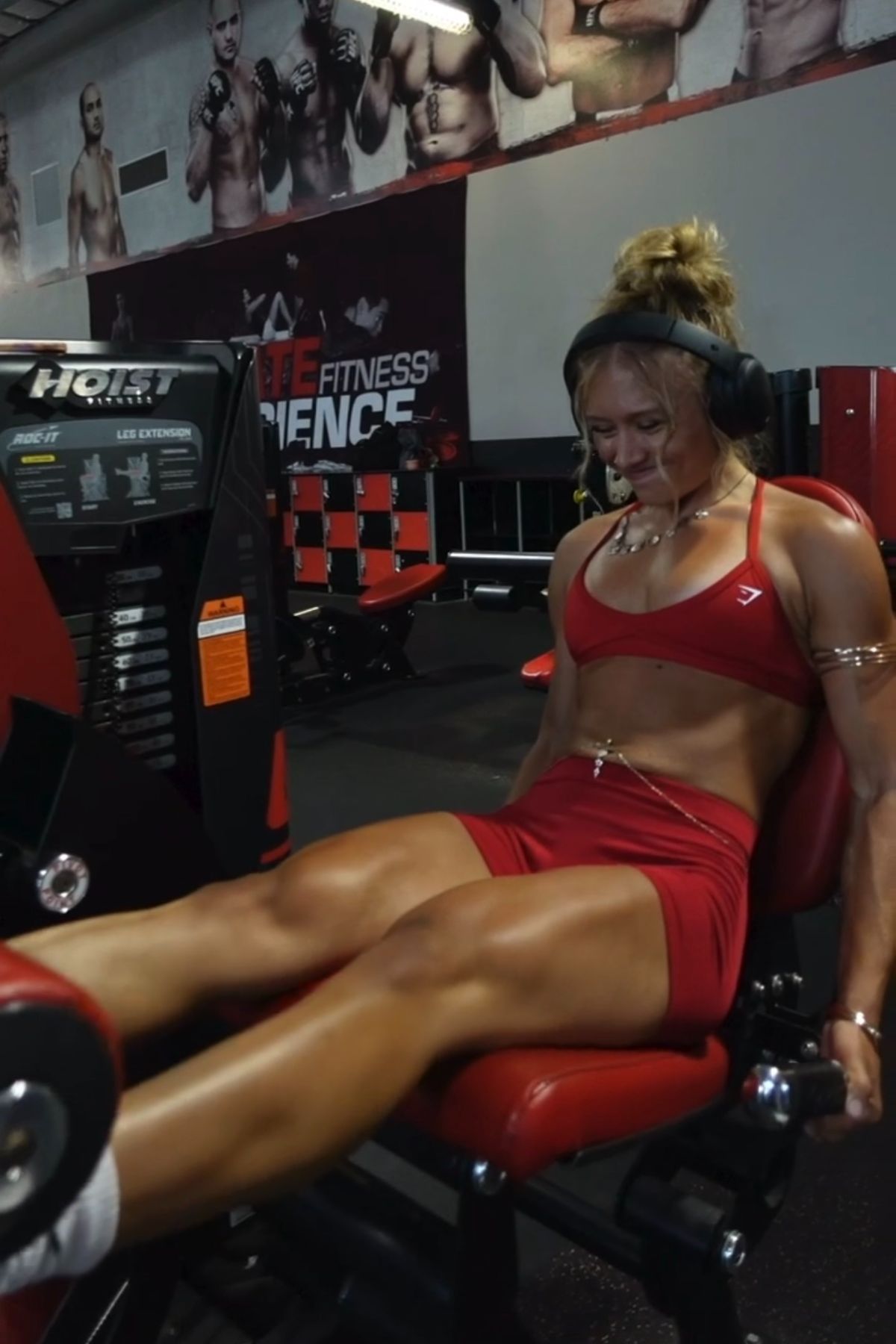 The coach wearing a red gym set and headphones, working out on a leg extension machine.