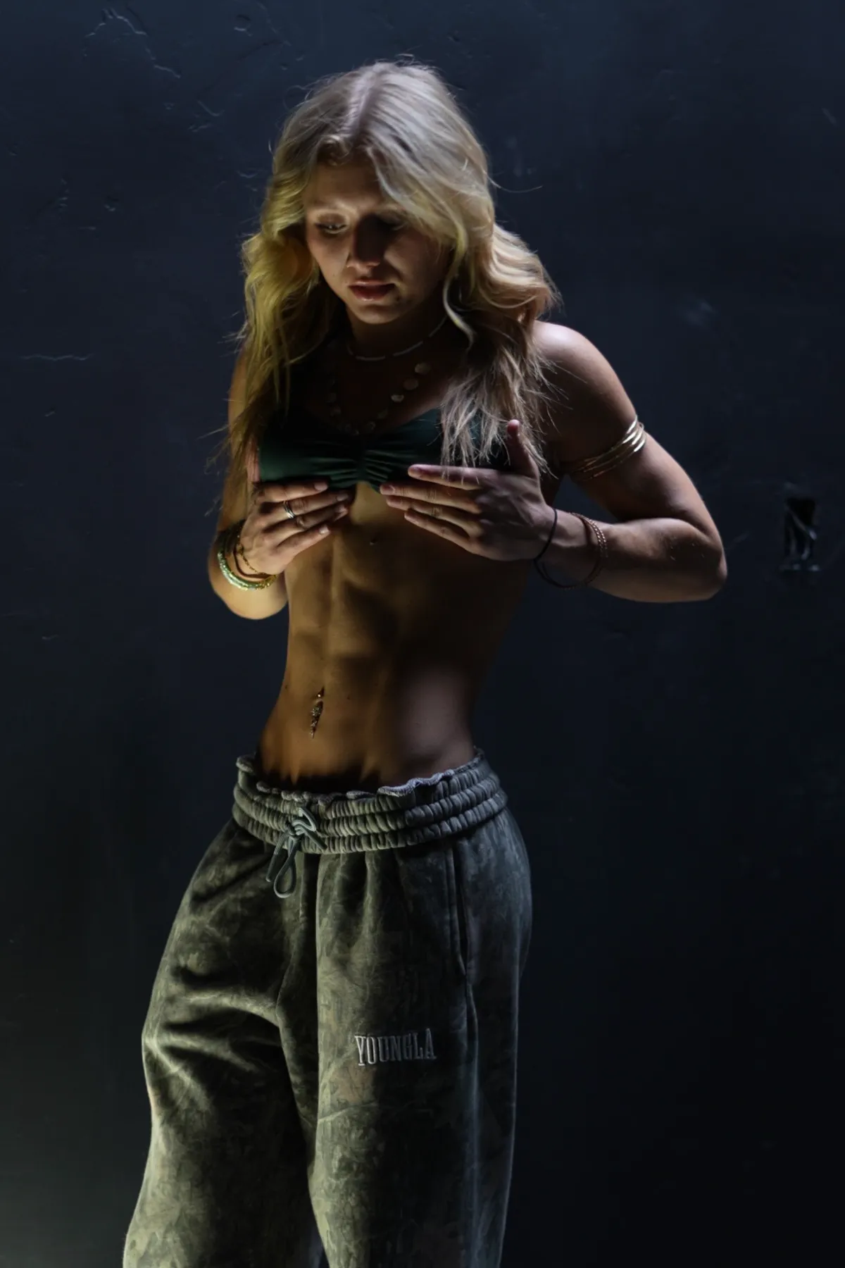 The coach showing her defined abs in a dark room, wearing a top and baggy sweatpants.